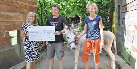 Stadtwerke-Geschäftsführerin Andrea Vogt überreicht Simon Brücken, dem Leiter des Abenteuerspielplatzes Friedrich-Wilhelms-Hütte, einen Spendenscheck in Höhe von 801,20 Euro. Maria Gerdes, ebenfalls Mitarbeiterin der Freizeiteinrichtung und für den Großtierbereich zuständig, hält Esel Felix an der Leine, der künftig die Kutsche ziehen wird.